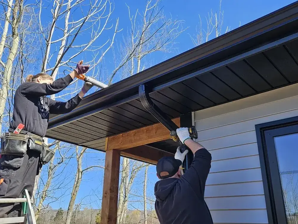 Two Wildawbren installers mounting a black seamless gutter along the gable of a beige lap-sided home, with cedar porch posts below