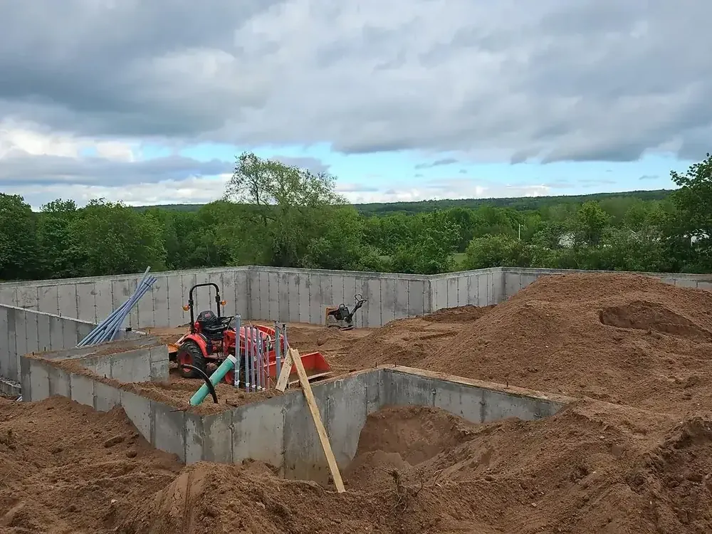 A Wildawbren new build at the foundation stage: poured concrete walls, excavated terrain, a compact Kioti tractor on the pad, and open Nova Scotia countryside beyond