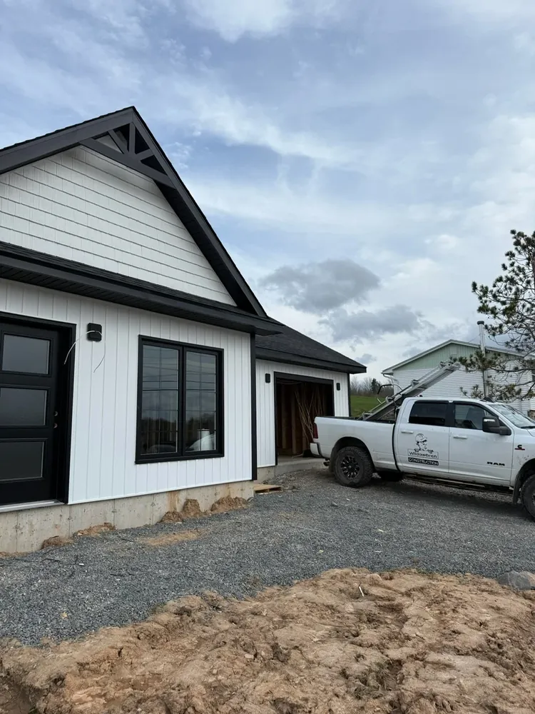 A Wildawbren branded GMC pickup parked beside a freshly finished white-sided home with black trim, fresh gravel still around the foundation