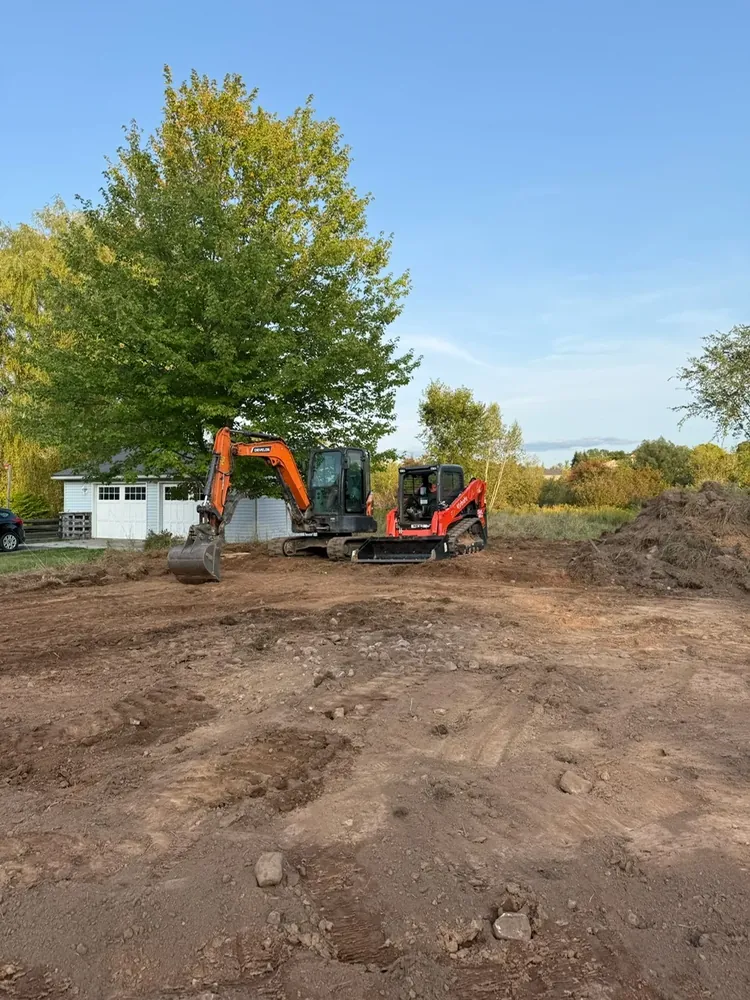 A Wildawbren mini excavator parked mid-dig on a residential lot in autumn, fresh dirt mounded behind it and a yellow-leafed tree above
