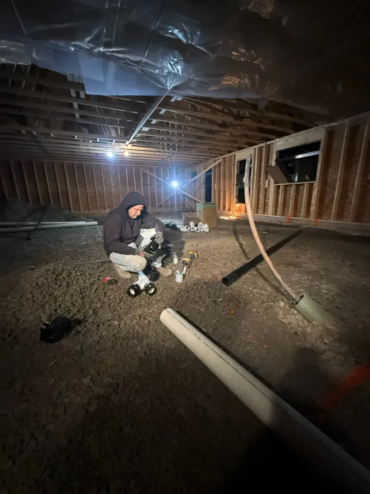 A Wildawbren tradesperson kneeling in a vapour-sealed basement, fitting a plumbing rough-in under the floor joists by work-light